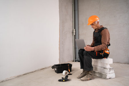 builder or repairman senior man in safety helmet sitting using laptop computer ordering online on...