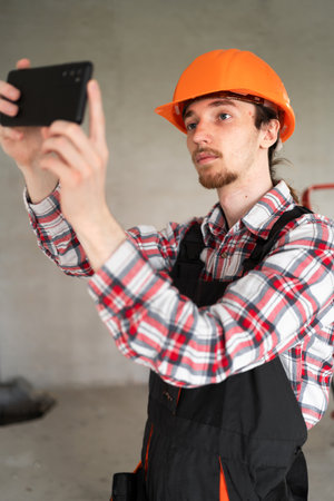 Builder, foreman, repairman wearing tool belt hard hat and overalls at construction site make photo by smartphone.の写真素材