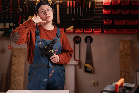 Woman joiner wears workwear in workshop scene with tools hanging on wall showing phone gesture say call me. female carpenter late evening in authentic garage.の写真素材