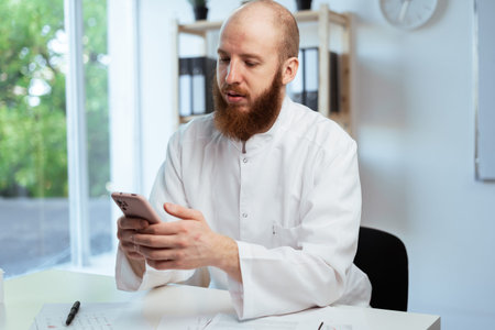 Bearded male doctor wearing white coat sitting at desk working with smartphone in clinic setting