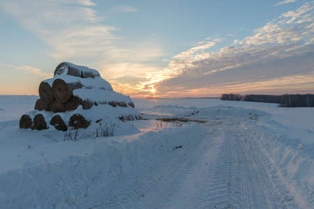 winter landscape with view of snow-covered straw bales in field at sunsetの写真素材