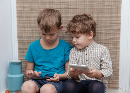 two brothers of European appearance four and seven years old enjoys playing games on your tablet and smartphone , sitting on the windowsillの写真素材