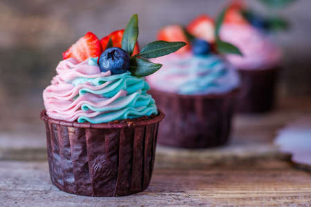 beautiful homemade cupcakes with pink and turquoise curd cream, decorated with strawberries and blueberries, on a wooden tableの写真素材