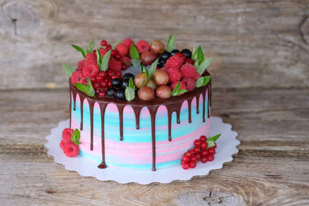 beautiful homemade summer cake with fresh raspberries, currants, gooseberries, decorated with chocolate icing, blue and pink cheese cream, on a wooden backgroundの写真素材