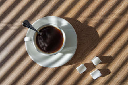 Hot coffee in a white cup with a saucer and three pieces of sugar on the office table near the window with blindsの写真素材