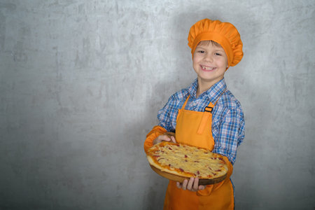 European boy dressed as a cook holds a freshly prepared homemade pizza against a gray wall. harvesting for advertising pizzaの写真素材
