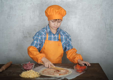 European boy dressed as a cook is engaged in cooking homemade pizza. rolls out the dough, lays out the sausage and tomatoesの写真素材