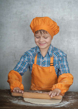 European boy dressed as a cook is engaged in cooking homemade pizza. rolls out the dough, lays out the sausage and tomatoesの写真素材
