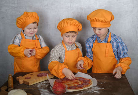 three cute European boys in costumes chefs enthusiastically busy cooking pizza. three brothers help mom to cook pizza with sausage and cheeseの写真素材