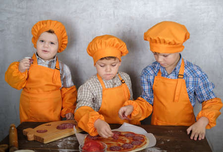 three cute European boys in costumes chefs enthusiastically busy cooking pizza. three brothers help mom to cook pizza with sausage and cheeseの写真素材