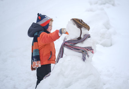 a little boy inserts a carrot instead of a nose to a snowman in winter for a walkの写真素材