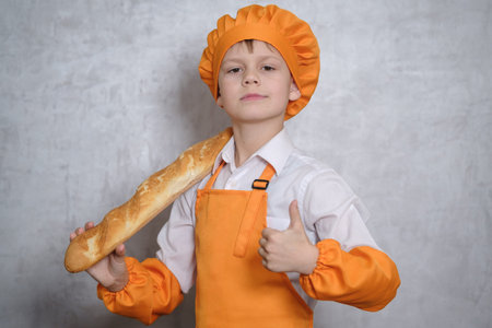 a cute boy in an orange cook suit holds a baguette and shows a like gesture on a gray wall backgroundの写真素材