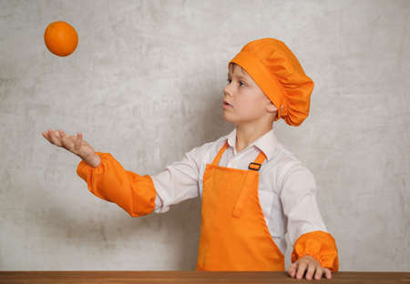 cute European boy in an orange suit chef tosses an orange up standing at a table on the background of the gray wallの写真素材
