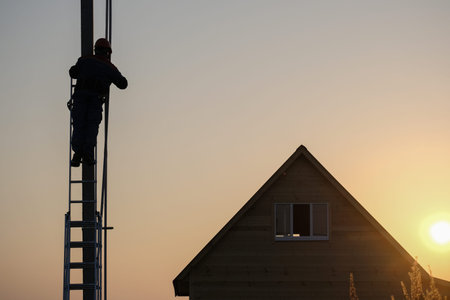 electrician performs installation of electrical equipment on the power line support near a wooden house in the evening at sunset. electrification of the new houseの写真素材