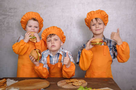 three cute boys in costumes cooks engaged in cooking homemade burgers. three brothers preparing family dinnerの写真素材