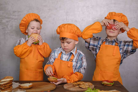 three cute boys in costumes cooks engaged in cooking homemade burgers. three brothers preparing family dinnerの写真素材