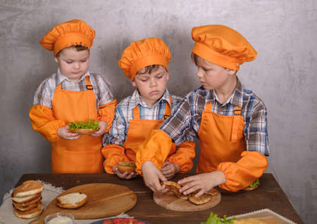 three cute boys in costumes cooks engaged in cooking homemade burgers. three brothers preparing family dinnerの写真素材