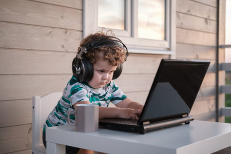 funny cute boy with curly hair in big headphones sitting at a laptop on the terrace of the house on...