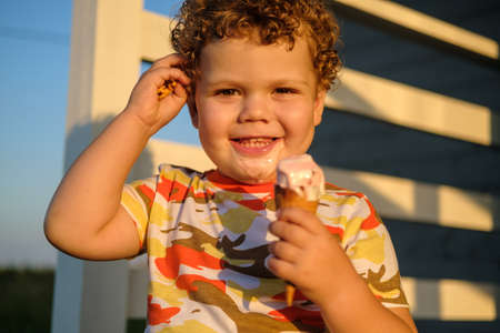 cute curly haired boy eating ice cream waffle cone outside on a summer Sunny dayの写真素材