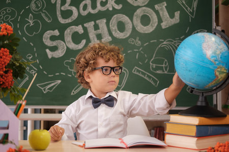 a cute curly-haired boy is sitting at a table against the background of a blackboard with the inscription back to schoolの写真素材