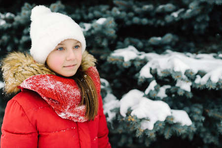 portrait of a girl in a red jacket, near the green tree on the street in the winter in the snowの写真素材