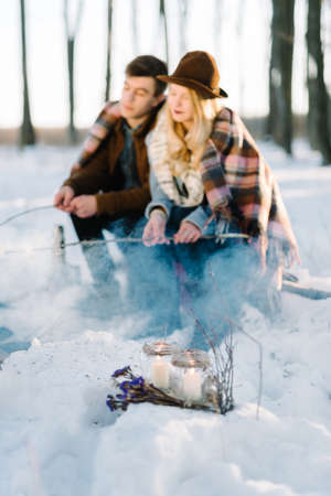 hipster stylishly dressed young couple in a warm blanket, sit by the fire in the winter forestの写真素材