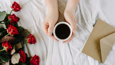 woman's hand, a cup of black coffee and a bouquet of red roses, envelope with congratulations on a white backgroundの写真素材