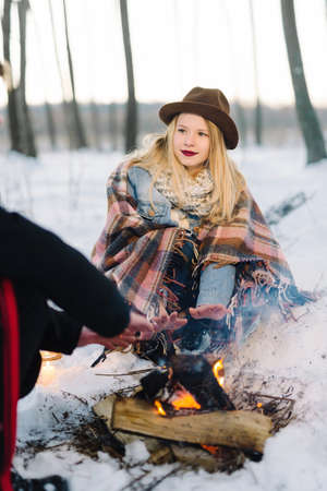hipster stylishly dressed young couple in a warm blanket, sit by the fire in the winter forestの写真素材