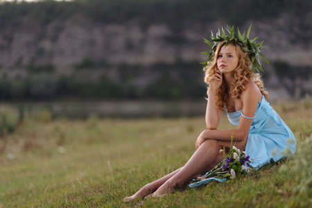 Girl in a blue dress on her head wreath made from tree branches, on a green lawn against a background of rocksの写真素材