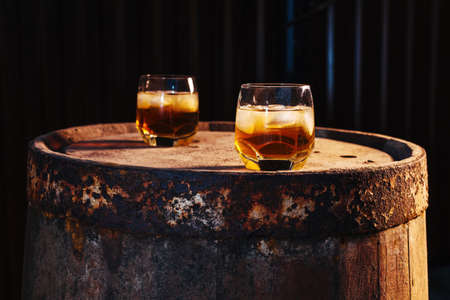 A glass of whiskey with ice on a wooden background close-up, in the background of a barrel of whiskeyの写真素材