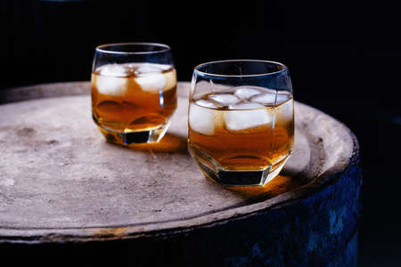 A glass of whiskey with ice on a wooden background close-up, in the background of a barrel of whiskeyの写真素材