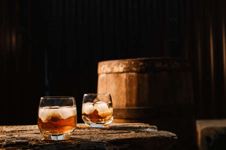 A glass of whiskey with ice on a wooden background close-up, in the background of a barrel of whiskeyの写真素材