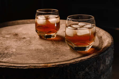 A glass of whiskey with ice on a wooden background close-up, in the background of a barrel of whiskeyの写真素材