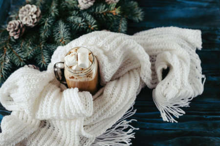 New Year's cappuccino with marshmallows, white knitting scarf on an old blue wooden table, for decoration, pine for a branch with cones, space for textの写真素材