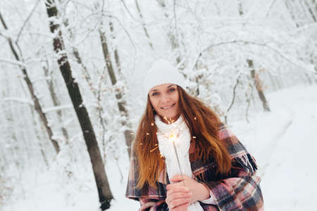 A woman holding a bright festive Christmas spark in her hand, in a white hat, wrapped in limbs, in a snow-covered winter forestの写真素材