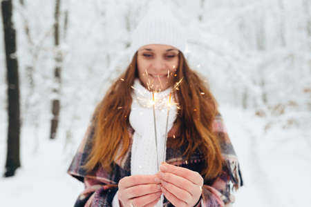 A woman holding a bright festive Christmas spark in her hand, in a white hat, wrapped in limbs, in a snow-covered winter forestの写真素材