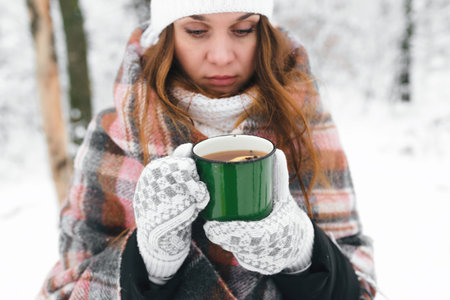 Girl in a white hat wrapped in a blanket, holding a green mug of tea in winter, close-up portrait