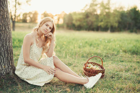 romantic girl in a dress near the tree with a basket of apples, in the setting sun of the summer sunの写真素材