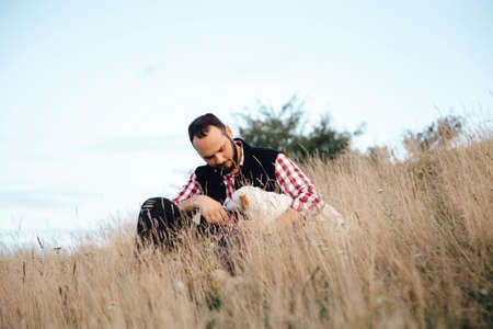A man wearing a dark vest and jeans sitting on the ground and playing outdoors in the park with his dog. The young owner hugs his pet.の写真素材