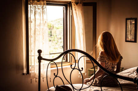 Long-haired woman sitting on the bed and looking out the windowの写真素材