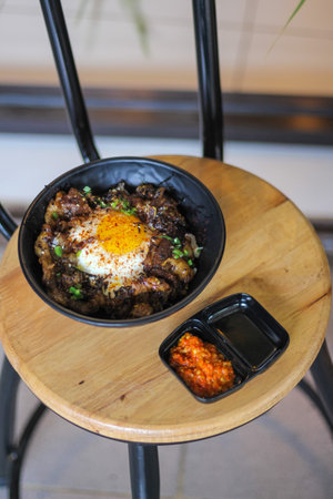 steamed rice with beef and fried egg in black bowl on wooden tableの写真素材