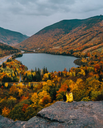 Artist Bluff Trail New Hampshire Adventure Girl in Yellow Coat Sits on Edge of Mountain Beautiful Foliage Autumn Scenery Lake Moody Fall Vibes.の写真素材