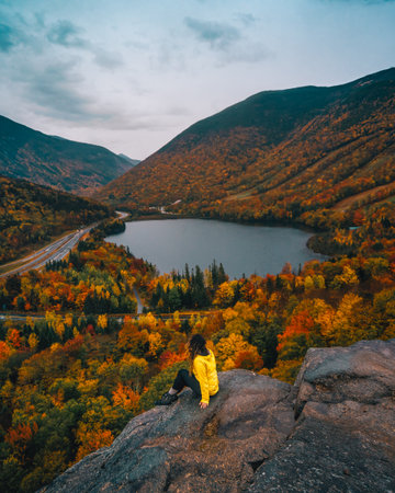 Autumn Female Adventure Hiker Sitting on Edge of Mountain Epic View From Artist Bluff Trail New Hampshire Beautiful Foliage Scenery Lake Yellow Jacketの写真素材