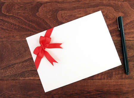 white envelope of greeting card with red ribbon bow and black pen on dark brown wooden table floor, flat lay close-up top viewの写真素材