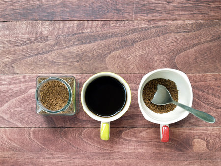 soluble instant coffee powder in transparent glass jar and one cup of black coffee and prepare to brew another cup with silver teaspoon on dark brown wooden table, close up top view with copy spaceの写真素材