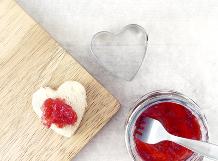 strawberry jam spread on small heart shape white bread on cutting board, pastry cutter and berry jam in glass jar with silver fork on blotting paper, wedding appetizer menu, flat lay close up top viewの写真素材