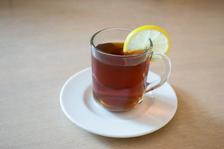 Glass cup of tea with lemon on a white saucer on a wood table.