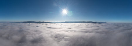 An aerial panoramic view captures a vast sea of white clouds stretching below mountain peaks,...