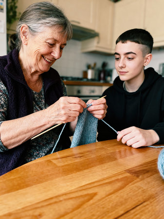 A smiling grandmother patiently instructs her grandchild in knitting, sharing traditional skills at a home kitchen table.の素材
