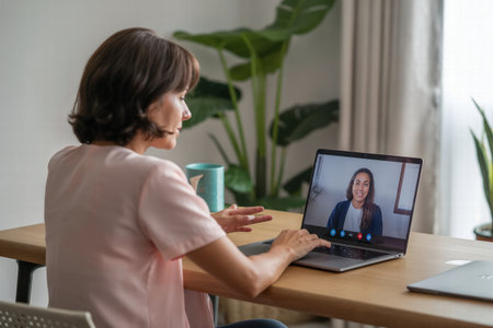 An adult woman sits at a wooden desk, engaged in a video conference on her laptop with a remote colleague.の素材